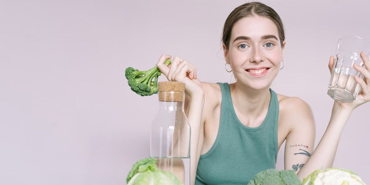 Woman in tank top smiling with broccoli, cabbage, and glass of water on table.