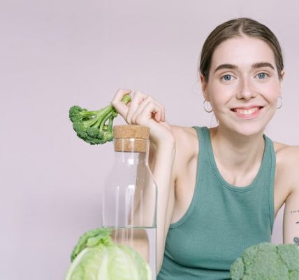 Woman in tank top smiling with broccoli, cabbage, and glass of water on table.