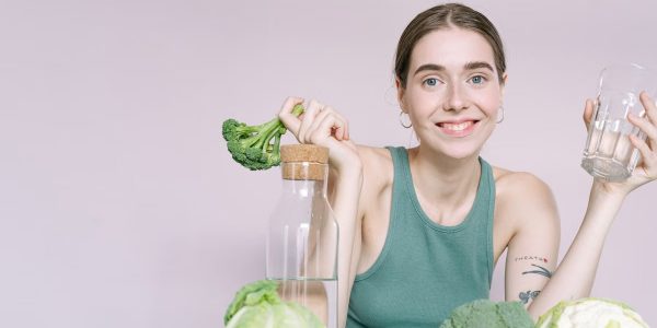 Woman in tank top smiling with broccoli, cabbage, and glass of water on table.