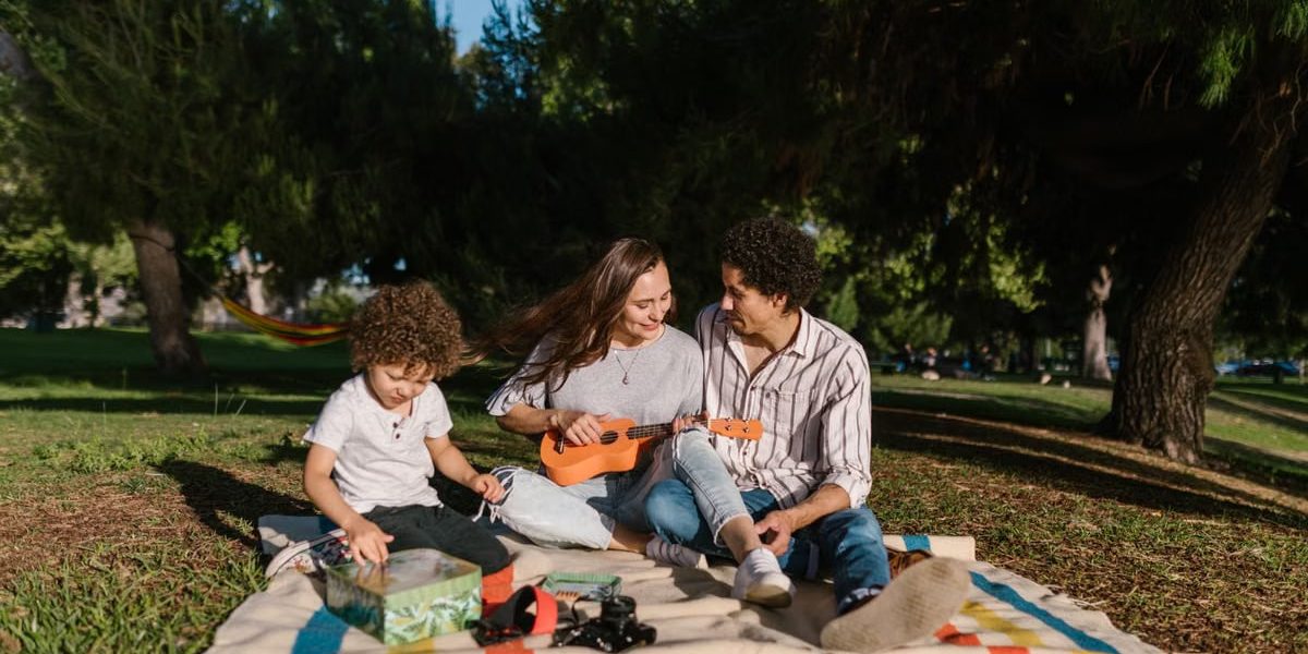 A joyful family enjoying a summer picnic with a ukulele in a sunny park setting.